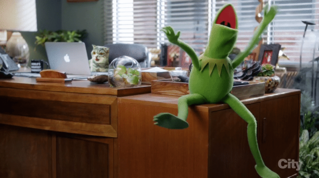 Kermit the frog sitting at the corner of his desk waving his hands over his head in joy
