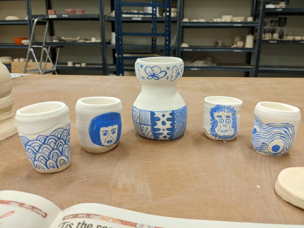 The homemade sake set before being glazed. Features the illustrations. From left - Japanese Sea waves, Yayoi Kusama, carafe patterns, a monkey, and ramen with eggs.