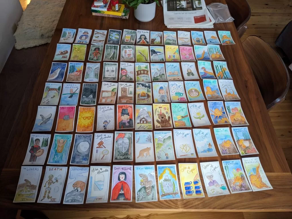 The original art of the Playful Beings tarot deck laid out in rows on a wooden table. This was taken before edits and some cards are now different.